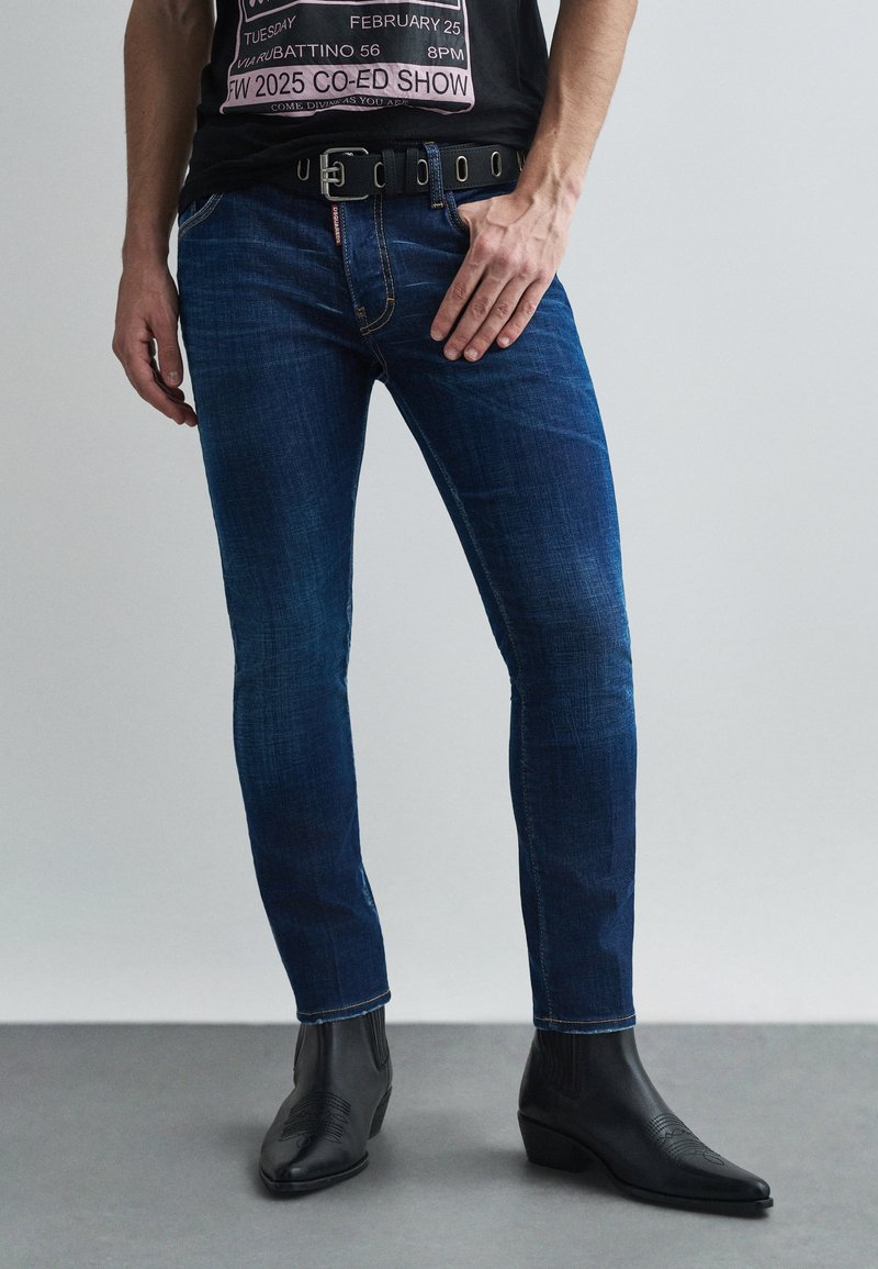 Vaqueros de denim azul oscuro ajustados con un ligero desgaste, que presentan un cinturón con acentos circulares, combinados con botas de cowboy negras de punta.