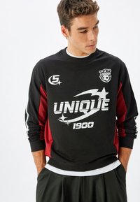Sudadera negra y roja de mangas largas, con un estampado gráfico blanco "UNIQUE 1900" y un logotipo en el pecho. Puños y dobladillo de canalé.
