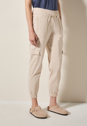 Persona che indossa pantaloni cargo jogger beige con polsini elasticizzati e un top blu navy, abbinati a sandali beige slip-on con fibbie.