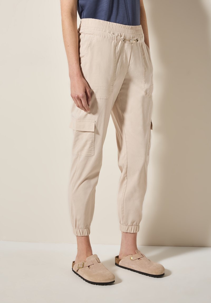 Personne portant un pantalon jogger cargo beige avec des poignets élastiques et un haut bleu marine, assorti à des sandales beige à enfiler avec des boucles.