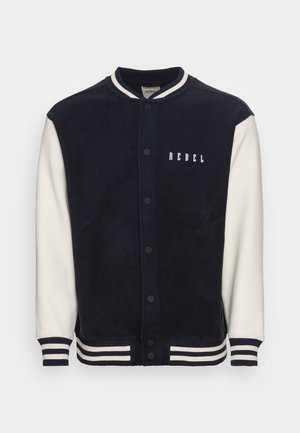 Veste universitaire bleu marine et blanche avec poignets et col rayés, boutons pression à l’avant, et le mot "REBEL" imprimé sur la poitrine gauche.