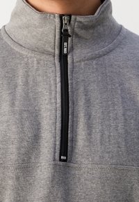 Gros plan d'une personne portant un sweat-shirt gris à fermeture éclair quart de zip avec un curseur de zip noir marqué "EST. 1993."