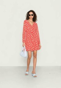 Mulher com cabelo encaracolado a usar óculos de sol redondos e um vestido longo de mangas compridas com padrão floral vermelho, segurando uma mala branca e usando saltos abertos de cor azul-clara.