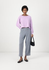 Pulover scurt de lavandă cu un decolteu larg în formă de barcă, purtat împreună cu pantaloni gri cu picioare largi. Modelul ține o mică geantă neagră și poartă balerini negri.
