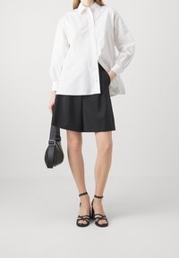 Frau trägt ein weißes Hemd mit Knöpfen, schwarze knielange Shorts, schwarze Absatzsandalen und hält eine schwarze Handtasche mit breitem Trageriemen.