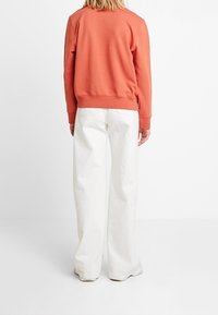 Felpa arancione con scollo rotondo e polsini a costine, abbinata a pantaloni bianchi a gamba larga. Il tessuto sembra morbido con una vestibilità comoda.