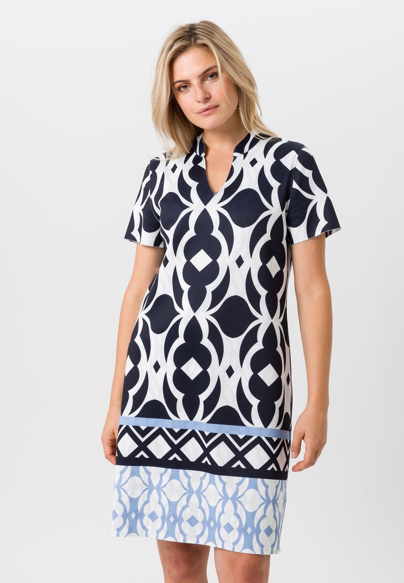 Korte mouw jurk met een marineblauw en witte geometrische print en een V-hals. Het ontwerp bevat gelaagde lichtblauwe accenten aan de zoom.