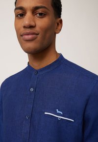 Blauwe linnen shirt met een bandkraag, knoopsluiting en geborduurd logo op de borstzak, met een gestreepte acccent op de zak.
