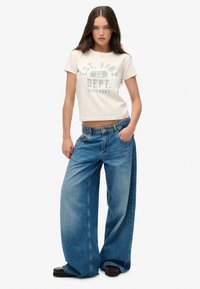 Afskåret cremefarvet t-shirt med blå tekst, parret med brede lyseblå jeans. Modellen står med den ene hånd i lommen, og sorte sko fuldender looket.