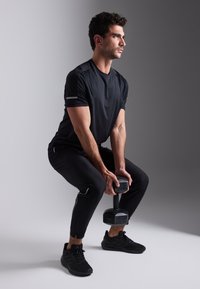Een man in een zwart sportshirt en strakke zwarte broek houdt een dumbbell vast. Hij draagt zwarte sneakers en heeft een neutrale houding tegen een grijze achtergrond.