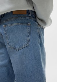 Světle modré džíny z denimu mají zadní kapsu s prošitými detaily a na pásku koženou nášivku v barvě tan s nápisem „DENIM GOODS“.