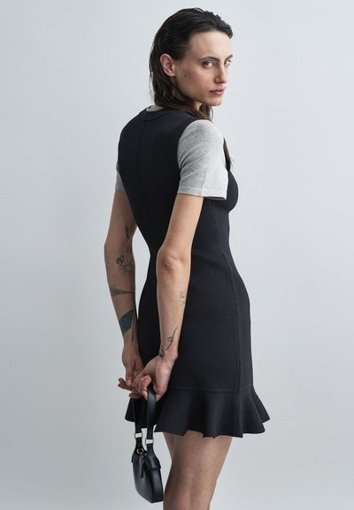 Robe noire ajustée avec des manches courtes grises, en tissu texturé et avec un ourlet évasé. La mannequin tient un petit sac à main noir. Design simple et moderne.