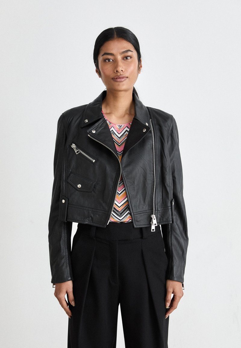Veste en cuir noir de motard coupée avec des accessoires en métal argenté, poches zippées et une finition texturée, portée par-dessus un haut à motifs colorés.