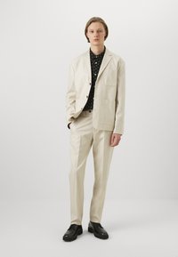 Ljusbeige suit med två fickor på jackan, bärs över en svart skjorta med vita prickar, tillsammans med svarta skor.