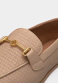 Mocassin beige avec texture tissée et détail en métal doré en forme de mors de cheval sur la partie supérieure, surpiqûres visibles et semelle lisse.