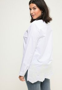 Camicia bianca a maniche lunghe con colletto a punta e orlo ricamato a scollatura. Tessuto morbido con una texture liscia. Abbinata a jeans blu.