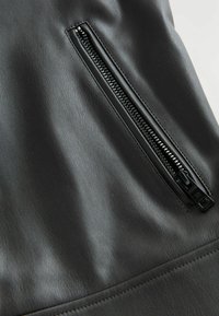 Černá umělá kůže s bočním detailem na zip. Textura je hladká, s jemným leskem a kontrastním stehem podél lemu.