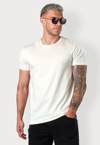 Beige Kurzarm-T-Shirt mit Rundhalsausschnitt und minimaler Markenkennzeichnung. Es hat eine glatte Textur und eine schmale Passform, kombiniert mit dunklen Jeans.