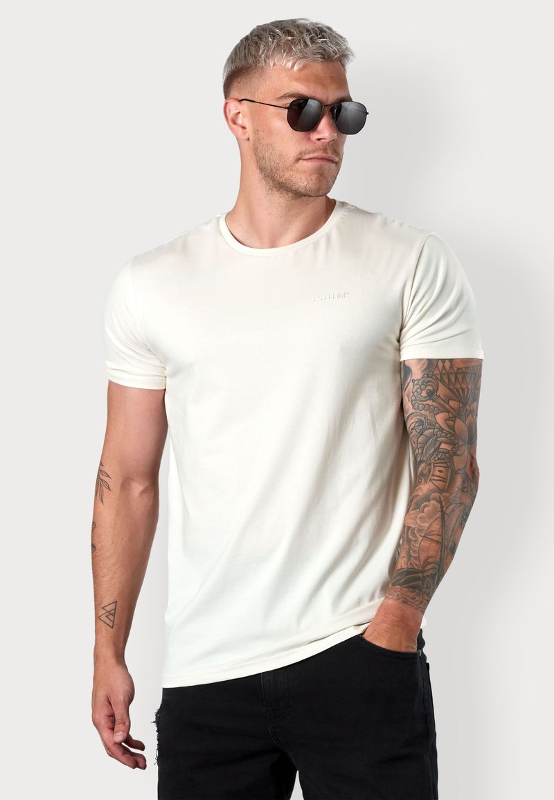 Beige Kurzarm-T-Shirt mit Rundhalsausschnitt und minimaler Markenkennzeichnung. Es hat eine glatte Textur und eine schmale Passform, kombiniert mit dunklen Jeans.