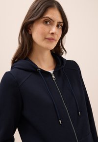 Haut de survêtement bleu marine avec fermeture éclair, cordon de serrage et deux poches. Présente un tissu texturé et une coupe décontractée, mettant en avant un design simple.