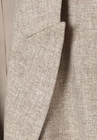 Solapa de blazer de tela tejida en beige y marrón claro que muestra textura y patrón con una ligera sombra en los pliegues de la tela.