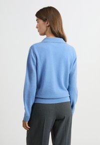 Jasnoniebieski wełniany sweter z kołnierzykiem, luźny krój i ściągacz na dole, zestawiony z szarymi, dopasowanymi spodniami. Gładka tekstura, minimalne detale.