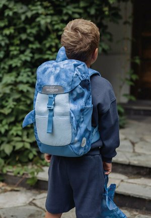 Child with short hair wearing a blue shark-themed backpack and matching lunch bag, standing outside near leafy greenery and stone steps.