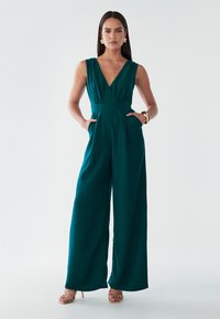 Teal ærmeløs jumpsuit med dyb V-hals, tætsiddende talje, sidelommer, brede ben og glat stof; parret med gyldne strop sandaler.