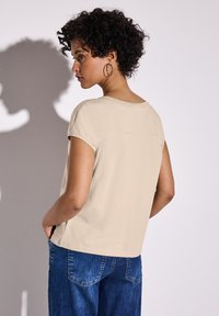 Beige top s krátkým rukávem a kulatým výstřihem, s detailem zadního švu. Kombinováno s tmavě modrými džíny. Hladká textura.