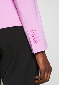 Růžová sako rukáv s čtyřmi ladícími knoflíky a hladkou texturou, doplněné černými kalhotami. Detailní záběr.