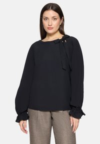 Zwarte blouse met een ronde halslijn, strikdetail bij de schouder en lange pofmouwen met gerimpelde manchetten. Soepele stoftextuur.