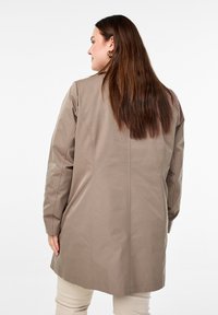 Pitkä beige takki, jossa on sileä rakenne, suora leikkaus, olkasaumat ja sivupaneelit. Huomaamattomat tikkaus yksityiskohdat ovat näkyvissä.