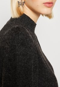 Blondynka z krótkimi włosami, ubrana w czarny, futrzany sweter z wysokim kołnierzem oraz srebrną kolczykiem w kształcie motyla w lewym uchu.