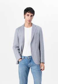 Blazer grigio chiaro e blu con motivo, colletto a becco, chiusura con un bottone e due tasche, abbinato a una t-shirt bianca e jeans blu.
