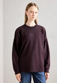 Sudadera púrpura oscura con cuello redondo, hombros caídos y logo bordado en el pecho; combinada con jeans azules.
