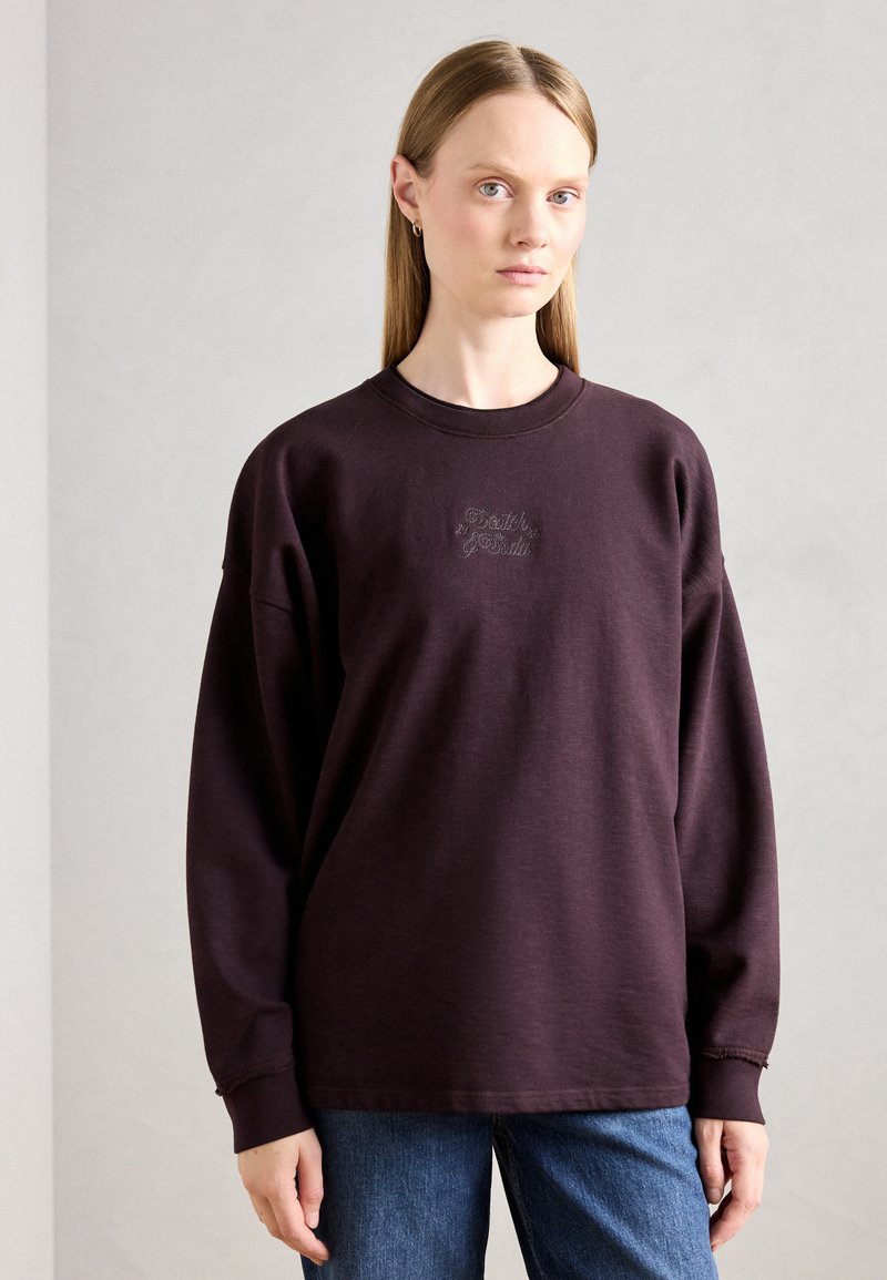 Sudadera púrpura oscura con cuello redondo, hombros caídos y logo bordado en el pecho; combinada con jeans azules.