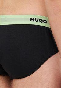Culottes en coton noir avec une ceinture élastique vert clair, arborant le logo "HUGO" en noir. Le design est lisse et ajusté.