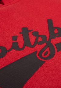 Rode hoodie met zwart gestructureerde letters en een driehoekige grafiek. De stof lijkt zacht met een lichte rek.