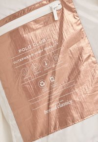 Metaliczna różowo-złota tkaninowa saszetka z białym suwakiem oraz nadrukowanym logo Polo Club, ikonami cech produktu i tekstem dotyczącym właściwości odzieży wierzchniej.