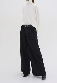 Biały sweter z golfem o luźnym kroju, w zestawieniu z czarnymi spodniami z wysokim stanem, wyposażonymi w pasek z prostokątną metalową klamrą.