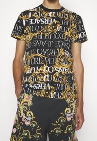 Svart t-shirt med gyllene barockmönster och vit text, med "Versace"-branding. Designen är djärv och kombinerar ornamentala inslag och kontrasterande färger.