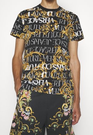 Camiseta negra con patrones barrocos dorados y texto blanco, que presenta la marca "Versace". El diseño es audaz, combinando acentos ornamentales y colores contrastantes.
