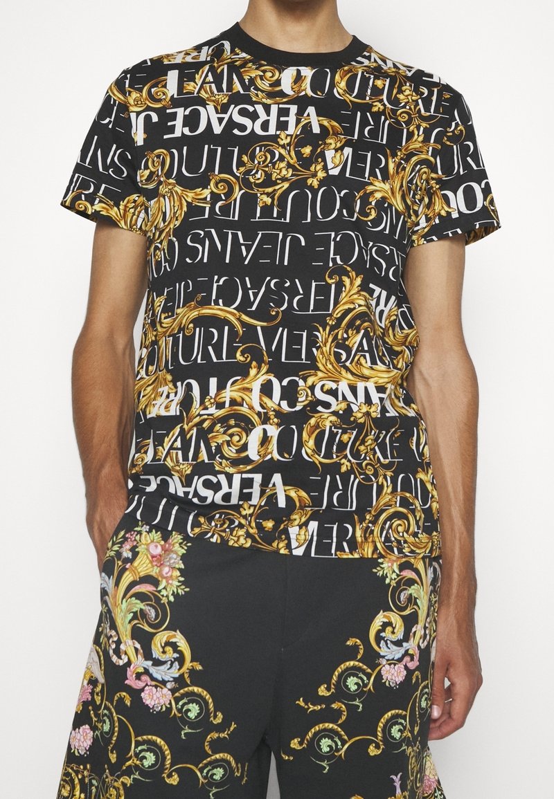 Svart t-shirt med gyllene barockmönster och vit text, med "Versace"-branding. Designen är djärv och kombinerar ornamentala inslag och kontrasterande färger.
