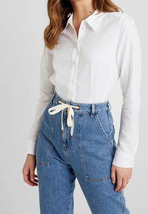 Kobieta ubrana w białą koszulę na guziki włożoną do wysokich niebieskich dżinsów z białym sznurkowym paskiem zawiązanym na kokardę.