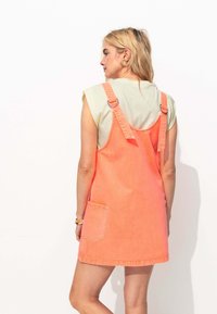Robe salopette en denim de couleur corail vif avec des bretelles ajustables, une poche latérale et une texture douce. Présente un design sans dos pour un look décontracté.