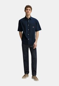 Camicia a maniche corte blu navy con taschino sul petto e un piccolo logo, abbinata a pantaloni scuri e sneakers beige. Tessuto liscio.