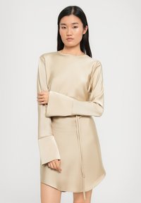 Beige satiini mekko, jossa on pitkät, leveät hihat, pyöreä pääntie ja kiristettävä vyötärö. Mekossa on sileä pinta ja hienovarainen kiilto.