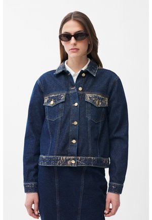 Junge Frau trägt eine dunkelblaue Jeansjacke mit abgenutzten goldenen Patches und einem passenden Rock, posiert mit schwarzer Sonnenbrille.