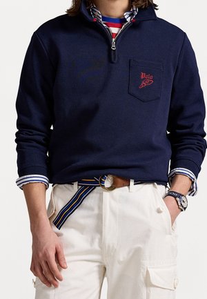 Uomo indossa un maglione blu navy con zip a collo alto, camicia a righe sotto, pantaloni cargo bianchi, cintura a righe e orologio al polso sinistro.