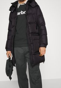 Svart pufferjacka med huva, med dragkedja framtill, flera fickor och quiltat mönster. Buren över en mörk tröja och byxor.